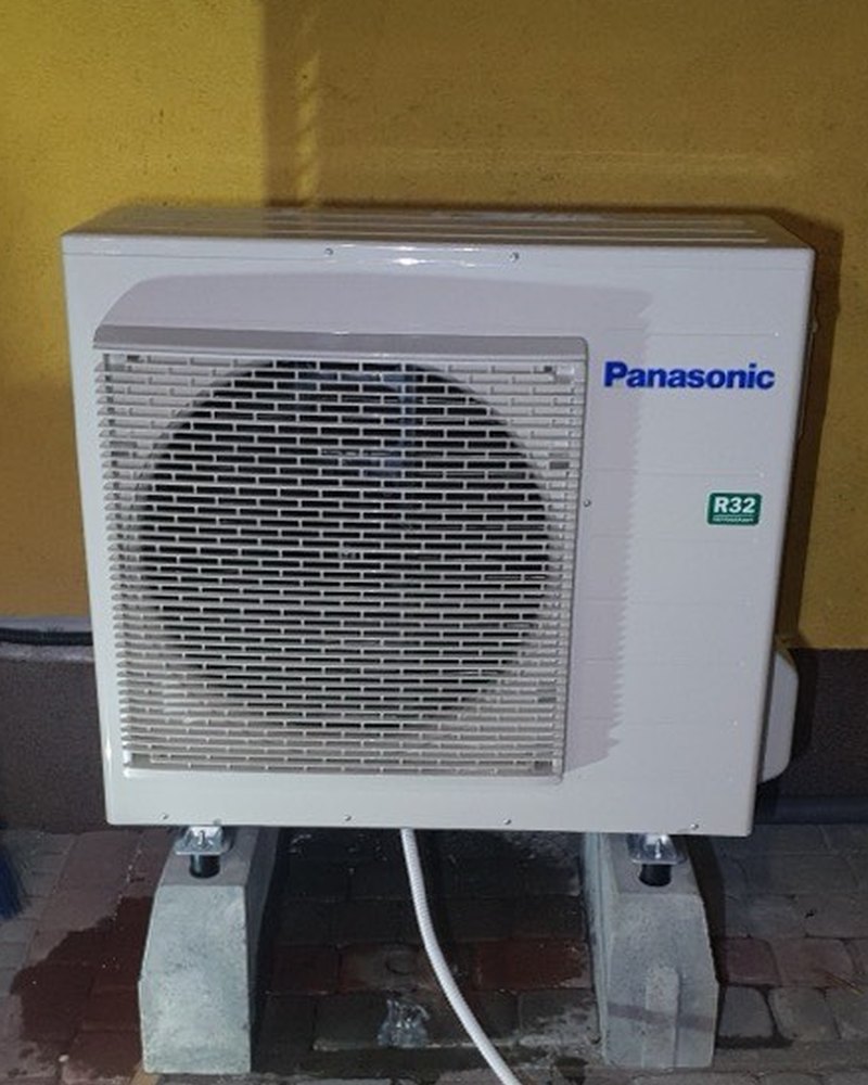 Jednostka zewnętrzna klimatyzacji Panasonic R32 na bloczkach przy musztardowej elewacji budynku — montaż Instal-Home